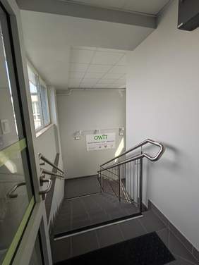Pokaż zdjęcie: Klatka schodowa wewnątrz nowoczesnego budynku. Schody wyłożone są ciemnymi płytkami, a po obu stronach znajdują się metalowe poręcze ze stalowymi elementami. Na ścianie naprzeciwko schodów wisi duży plakat z napisem "OWiT" oraz informacjami poniżej, co wskazuje na Ośrodek Wspierania i Testów. Ściany są jasnoszare, a sufit wykończony jest płytami sufitowymi. Po lewej stronie zdjęcia widać okno, przez które wpada naturalne światło, oświetlając klatkę schodową.
