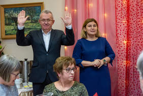 Pokaż zdjęcie: Mężczyzna w garniturze z uniesionymi rękami stoi obok kobiety w granatowej sukience na tle różowej dekoracji ze światełkami. W tle obraz przedstawiający krajobraz, a na pierwszym planie siedzące przy stole osoby.