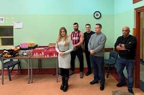 Pokaż zdjęcie: Na pierwszym planie przemawia kobieta, a w tle za nią kilkoro mężczyzn, w tym kulturysta, a na stoliku obok znajduja się paczki mikołajkowe