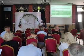 Pokaż zdjęcie: Grupa uczestników siedzi na czerwonych krzesłach w sali konferencyjnej Villa Tradycja, słuchając prezentacji kobiety siedzącej przy laptopie. Na ekranie wyświetlany jest slajd z katalogiem prac architektonicznych w Module E, przedstawiający kilka punktów w zielonych ramkach. Dekoracje w tle obejmują biało-zielony łuk kwiatowy z logo Villa Tradycja.