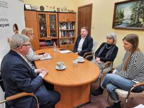 Pokaż zdjęcie: Przy owalnym stole siedzi pięć osób: trzy kobiety i dwóch mężczyzn. Rozmawiają ze sobą. Na stole leżą notesy, dokumenty, telefony, pieczątki i filiżanki. 