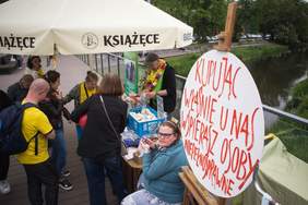 Pokaż zdjęcie: Przy stoisku stoją ludzie, na stole leżą przedmioty na sprzedaż. W prawym rogu zdjęcia napis kupując u nas wspierasz osoby niepełnosprawne.