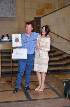 Pokaż zdjęcie: Uczestnik konkursu z dyplomem i prezentową torbą z logo PFRON, pozuje do fotografii z elegancką kobietą. Za nimi mównica z mikrofonami i marmurowe schody