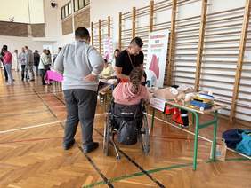 Pokaż zdjęcie: Osoba na wózku i mężczyzna stoją przy stanowisku z fantomami do nauki pierwszej pomocy w sali gimnastycznej, obok widoczny baner Środowiskowego Domu Samopomocy w Bytowie.