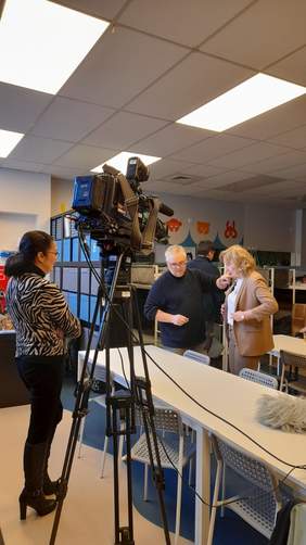 Pokaż zdjęcie: Przy stole stoi kobieta i mężczyzna, mężczyzna operator kamery wpina kobiecie mikrofon w klapę marynarki. Kobieta, blondynka ubrana jest w w beżowy garnitur i białą bluzkę. Parze przygląda się kobieta stojąca tyłem do zdjęcia, szatynka włosy związane w kucyk, ubrana w biało czarną marynarkę i czarne spodnie. Centralne miejsce na zdjęciu zajmuje kamera na statywie.