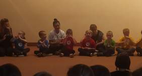 Pokaż zdjęcie: Na scenie po turecku siedzą przedszkolaki, na koszulkach maja kółeczka z narysowanymi buziami. Dzieci uczą się gestów służących komunikacji