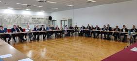 Pokaż zdjęcie: Duża grupa kobiet i mężczyzn siedzi w dużej sali przy stołach ustawionych w kształt litery „U”. Z tyłu sali, na ścianie widnieje napis: „sala konferencyjna”, jak również podwieszony jest obok głośnik.