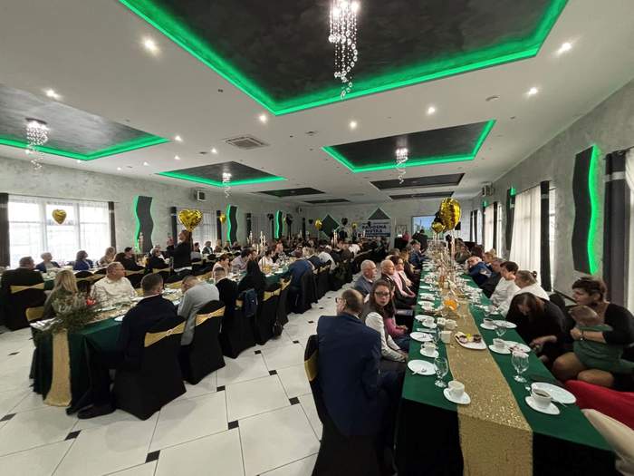 Pokaż zdjęcie: Grupa kilkudziesięciu osób siedzi w dużej sali przy stołach ustawionych w trzech rzędach. Na stołach przykrytych zielonym płótnem oraz złotym bieżnikiem po środku, ustawione zostały różne napoje i ciasta.