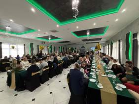 Pokaż zdjęcie: Grupa kilkudziesięciu osób siedzi w dużej sali przy stołach ustawionych w trzech rzędach. Na stołach przykrytych zielonym płótnem oraz złotym bieżnikiem po środku, ustawione zostały różne napoje i ciasta.