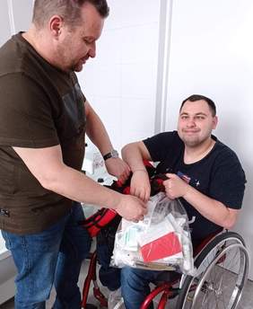 Pokaż zdjęcie: Mężczyzna siedzi na wózku inwalidzkim, drugi mężczyzna stoi przy wózku i podaje mu reklamówkę
