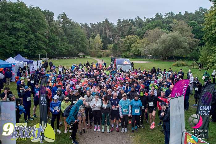 Pokaż zdjęcie: Duża grupa uczestników Biegu Pogoń Raka stoi na starcie w parku lub lesie, ubrana w sportowe stroje, wiele osób w kolorowych kurtkach i czapkach. W tle widoczny jest trawiasty teren rekreacyjny otoczony wysokimi drzewami oraz namioty organizacyjne.