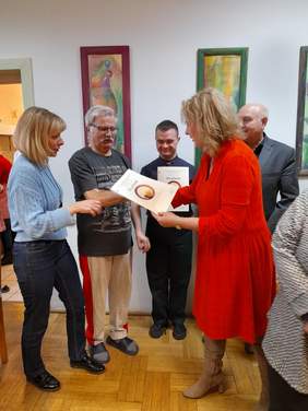 Pokaż zdjęcie: Kobieta, blondynka w czerwonej sukience wręcza dyplomy trzem osobom. 