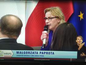 Pokaż zdjęcie: Na tle flagi Polski i Unii Europejskiej siedzi kobieta z mikrofonem w niebieskim garniturze, lekko przysłonięta głowami osób siedzących na widowni. Na dole zdjęcia napis Małgorzata Paprota Dyrektor Oddziału Lubelskiego PFRON, w prawym dolnym rogu zdjęcia widać kobiecą postać tłumacza języka migowego