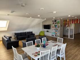 Pokaż zdjęcie: Nowoczesne, przytulne wnętrze z jadalnią i częścią wypoczynkową. Stół z białymi krzesłami, kanapy, regał z książkami i telewizor. Na ścianie kolorowe dekoracje, drewniana podłoga i skośny sufit z oknem dachowym.