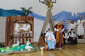 Pokaż zdjęcie: Na scenie siedzi kobieta z dzieckiem na ręku. Obok stoi mężczyzna, ma pochyloną głowę  z laską w dłoniach.Za nimi siedzą kobieta i mężczyzna.Za nimi stoją kobieta i mężczyzna przebrani za anioły.Po drugiej stronie szopka ze zwierzętami.