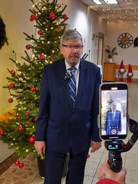 Pokaż zdjęcie: Na tle choinki stoi dyrektor Łódzkiego Oddziału PFRON Władysław Skwarka, który składa świąteczne życzenia. Jest nagrywany telefonem. Za nim widać bożonarodzeniowe dekoracje. 