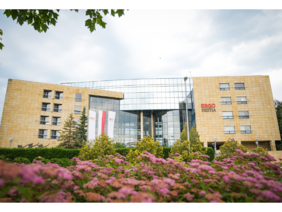 Pokaż zdjęcie: Budynek z kamienną elewacją i przeszklonym centralnym wejściem. Przed obiektem kwitnące różowe krzewy i zadbana zieleń.