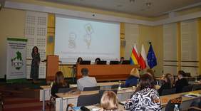 Pokaż zdjęcie: Zdjęcie przedstawia salę konferencyjną z uczestnikami siedzącymi przy stołach. Przy mównicy stoi kobieta w sukience, na ekranie wyświetlona jest prezentacja z napisem Wypożyczalnia technologii wspomagających dla osób z niepełnosprawnością. Po prawe stronie stoją trzy flagi: Polski, województwa podlaskiego i Unii Europejskiej. Po lewej stronie widoczny jest baner Regionalnego Ośrodka Polityki Społecznej w Białymstoku. Przy stole prezydialnym siedzą trzy osoby, korzystające z laptopów.