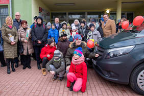 Pokaż zdjęcie: Przed budynkiem stoją zgromadzeni dorośli ludzie i dzieci po prawej stronie widać przód  samochodu marki bus . Auto jest ozdobione czerwonymi balonami.