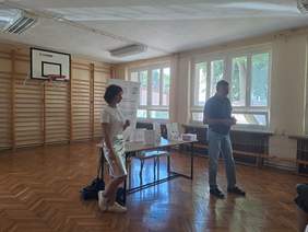 Pokaż zdjęcie: Przy stoliku na którym stoją torby z prezentami, katalogi i ulotki stoją dwie osoby, które rozmawiają z uczestnikami konferencji.