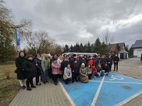 Pokaż zdjęcie: Duża grupa osób, w tym podopieczni i pracownicy Środowiskowego Domu Samopomocy w Sejnach, zgromadziła się na parkingu obok nowego, białego busa. Osoby ubrane są w ciepłe kurtki, co sugeruje chłodną pogodę. W tle widać domy, drzewa oraz znak parkingowy dla osób z niepełnosprawnościami.