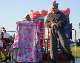 Pokaż zdjęcie: na scenie stoi osoba przebrana za wilka obok stoi osoba przebrana za babcię w ręku trzyma kołdrę. Jest to scena z bajki czerwony kapturek 