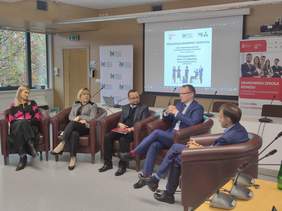 Pokaż zdjęcie: Mężczyźni i kobiety siedzą w fotelach i rozmawiają. Wsztscy sa ubrani elegancko. Fotele ułożone są w półkole.