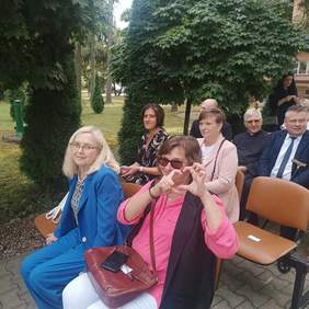 Pokaż zdjęcie: Zdjęcie zaproszonych gości siedzących na krzesełkach ustawionych w parku.