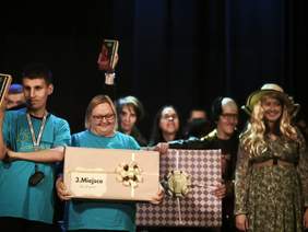Pokaż zdjęcie: Uczestnicy wydarzenia stoją na scenie, trzymając duże zapakowane pudełka z nagrodami. Osoba w środku trzyma karton z napisem „3. miejsce”. W tle widać innych uczestników oraz sceniczne oświetlenie.