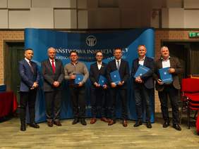 Pokaż zdjęcie: Grupa ośmiu mężczyzn w garniturach stoi na scenie podczas uroczystości, trzymają niebieskie teczki. Tło z logotypem „Państwowa Inspekcja Pracy – Okręgowy Inspektorat Pracy w Gdańsku”.