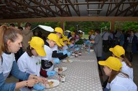 Pokaż zdjęcie: Przy długim stole siedzą dzieci. Jedzą naleśniki. Większość ma na głowach żółte czapeczki. 