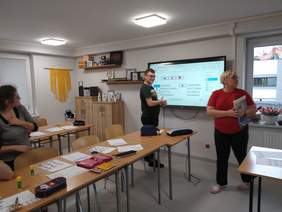 Pokaż zdjęcie: Przy tablicy interaktywnej nastolatek pokazuje napisy urządzeń biurowych. Na dalszym planie nauczycielka i inna uczennica.