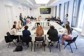 Pokaż zdjęcie: Ludzie siedzą przy kilku stołach ustawionych na planie kwadratu. Trwa dyskusja. Ludzie są zaangażowani w spotkanie. W dalszej perspektywie widać ekran z napisami na żywo.