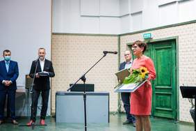 Pokaż zdjęcie: Przy mikrofonie stoi uśmiechnięta, krótko ścięta brunetka w okularach, pomarańczowej sukience i czerwono-czarnych butach na obcasie. W tle panowie w ciemnych garniturach, którzy wręczali statuetkę, kwiat słonecznika i dyplom oraz ogromne zielone