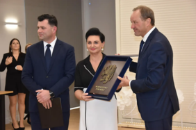 Pokaż zdjęcie: Trzy osoby w eleganckich strojach podczas uroczystości; kobieta w centrum trzyma ramę z herbem i napisem „Województwo Pomorskie”, mężczyzna po prawej wręcza nagrodę, drugi po lewej stoi z teczką, w tle goście.