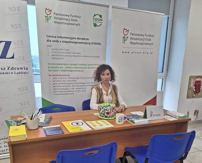 Pokaż zdjęcie: Za stołem na ktorym leża ulotki, siedzi młoda kobieta, przy stoliku stoją dwa krzesełka dla petentów. W tle za kobieta stoją dwa roll-upy z logo PFRON oraz CIDON.