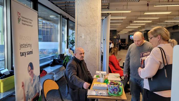 Pokaż zdjęcie: Przy stole w holu uniwersytetu stoją stoliki na których leżą ulotki i broszury. Przy stolikach siedzą eksperci, którzy odpowiadają na pytania.