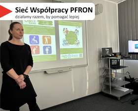 W centralnej części slajdu widać kobietę w czarnym stroju, stojącą przy tablicy projektowej i mówiącą do grupy. W tle jest wyświetlana prezentacja na tablicy. Po prawej stronie sali stoi wózek ze sprzętem i monitor, a przy oknie są zasłony. Na slajdzie w lewym górnym rogu jest biały pasek z czerwonym trójkątem i dużym napisem: „Sieć Współpracy PFRON”, a pod nim mniejszy: „działamy razem, by pomagać lepiej”.
