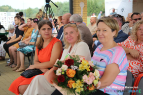 Pokaż zdjęcie: Zdjęcie trzech uśmiechniętych kobiet siedzących w pierwszym rzędzie. Jedna z nich trzyma kolorowy bukiet kwiatów.. 