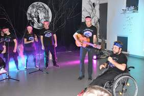 Pokaż zdjęcie: Na scenie oświetlonej w okolicy podłogi różowym światłem, w ciemnej scenerii trwa występ zespołu muzycznego. Frontman siedzi na wózku inwalidzkim, ma bandamke nagłowie, w tle stoją pozostali czlonkowie zespołu, jeden z nich gra na gitarze. Zespół ubramy jest w czarne podkoszulki z nazwa zespołu elektryczne mózgi. 