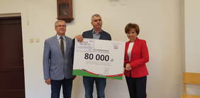 Pokaż zdjęcie: trzy osoby stoją obok siebie. Kobieta i mężczyzna trzymają dużą tablicę symbolizującą czek. 