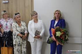 Pokaż zdjęcie: Na zdjęciu pod ścianą stoją 4 kobiety, jedna z nich trzyma kwiaty