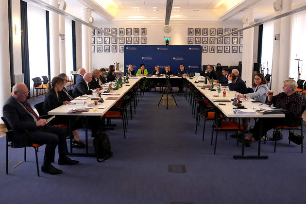 Ujęcie sali konferencyjnej podczas spotkania. Kilkanaście osób siedzi przy stołach ustawionych w podkowę. Na końcu sali znajduje się niebieska ścianka z identyfikacją wizualną instytucji, a na ścianach wiszą rzędy oprawionych fotografii.
