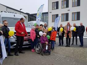 Pokaż zdjęcie: Grupa ludzi stoi przed srebrnym busem. Jedno z dzieci jest na wózku inwalidzkim. Uczestnicy trzymają balony i uśmiechają się. W tle nowoczesny budynek oraz windery z logo instytucji. 