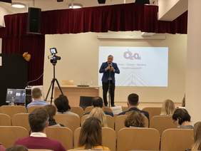 Pokaż zdjęcie: Na scenie z jasnego drewna stoi mężczyzna ubrany w granatowy garnitur białą koszulę i krawat. W ręku trzyma mikrofon. Z nim wyświetla się na ekranie napis OZON.