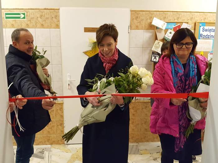 Pokaż zdjęcie: Dwie kobiety oraz jeden mężczyzna przecinają czerwoną wstęgę. Każda z tych osób trzyma w dłoniach bukiet róż. W tle widoczny jest korytarz wyłożony płytkami, zarówno na podłodze, jak i na ścianach. Za kobietą stojącą po środku, znajdują się drzwi od pracowni stolarsko-ślusarskiej. Po prawej stronie widoczne są natomiast dwie korkowe tablice ogłoszeniowe, na których przywieszone są różne kartki.