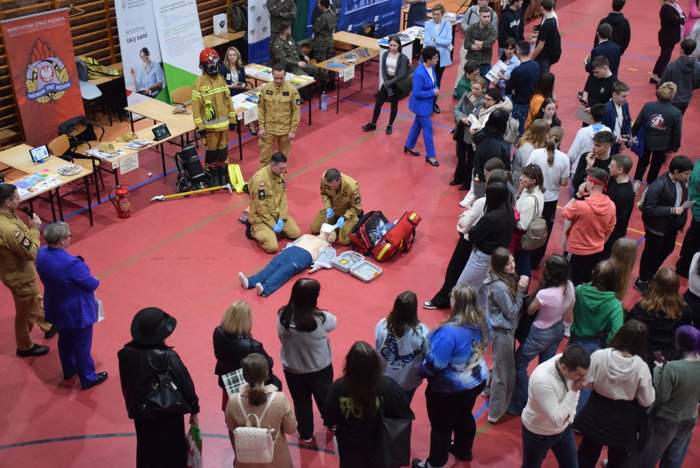 Pokaż zdjęcie: Grupa młodych osób zgromadzona jest w dużej hali sportowej i przygląda się szkoleniu z pierwszej pomocy, które prowadzą strażacy. W tle stoiska poszczególnych wystawców, w tym banery straży pożarnej oraz PFRON.