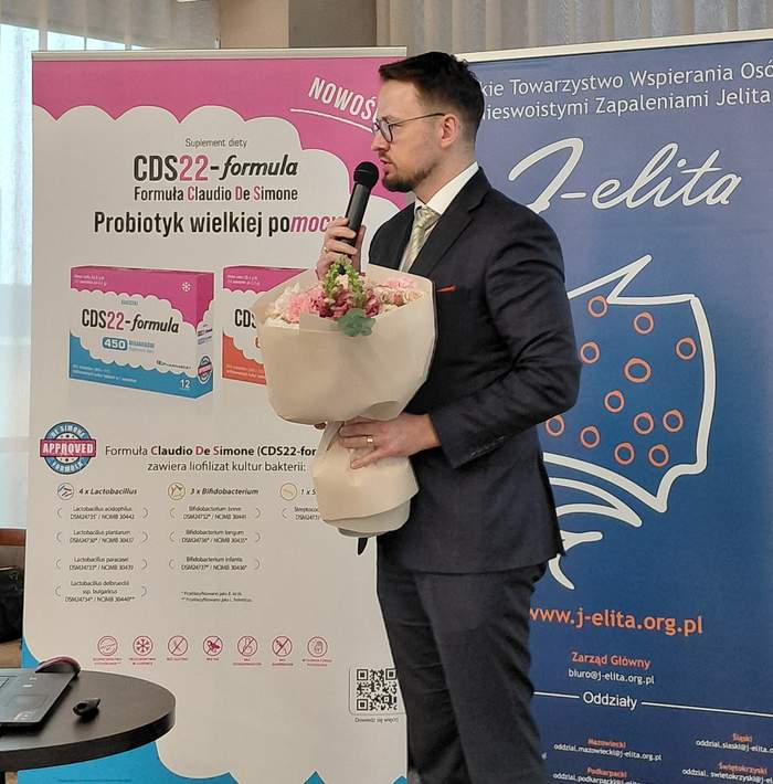Pokaż zdjęcie: Mężczyzna w garniturze i okularach przemawia do mikrofonu, trzymając w ramionach duży bukiet różowych kwiatów. Stoi przed tłem z logo i informacjami dotyczącymi suplementu diety CDS22-formula oraz logo i hasłami organizacji J-elita, Polskiego Towarzystwa Wspierania Osób z Nieswoistymi Zapaleniami Jelita.