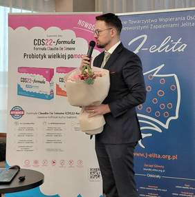 Pokaż zdjęcie: Mężczyzna w garniturze i okularach przemawia do mikrofonu, trzymając w ramionach duży bukiet różowych kwiatów. Stoi przed tłem z logo i informacjami dotyczącymi suplementu diety CDS22-formula oraz logo i hasłami organizacji J-elita, Polskiego Towarzystwa Wspierania Osób z Nieswoistymi Zapaleniami Jelita.