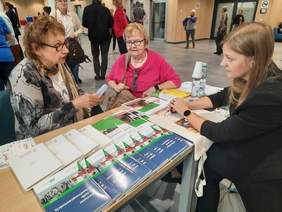 Pokaż zdjęcie: Obchodzony jest Dzień Seniora. Trzy panie siedzą przy stole i rozmawiają ze sobą. W tle widać spacerujących seniorów. Na stole leżą materiały promocyjne PFRON. 